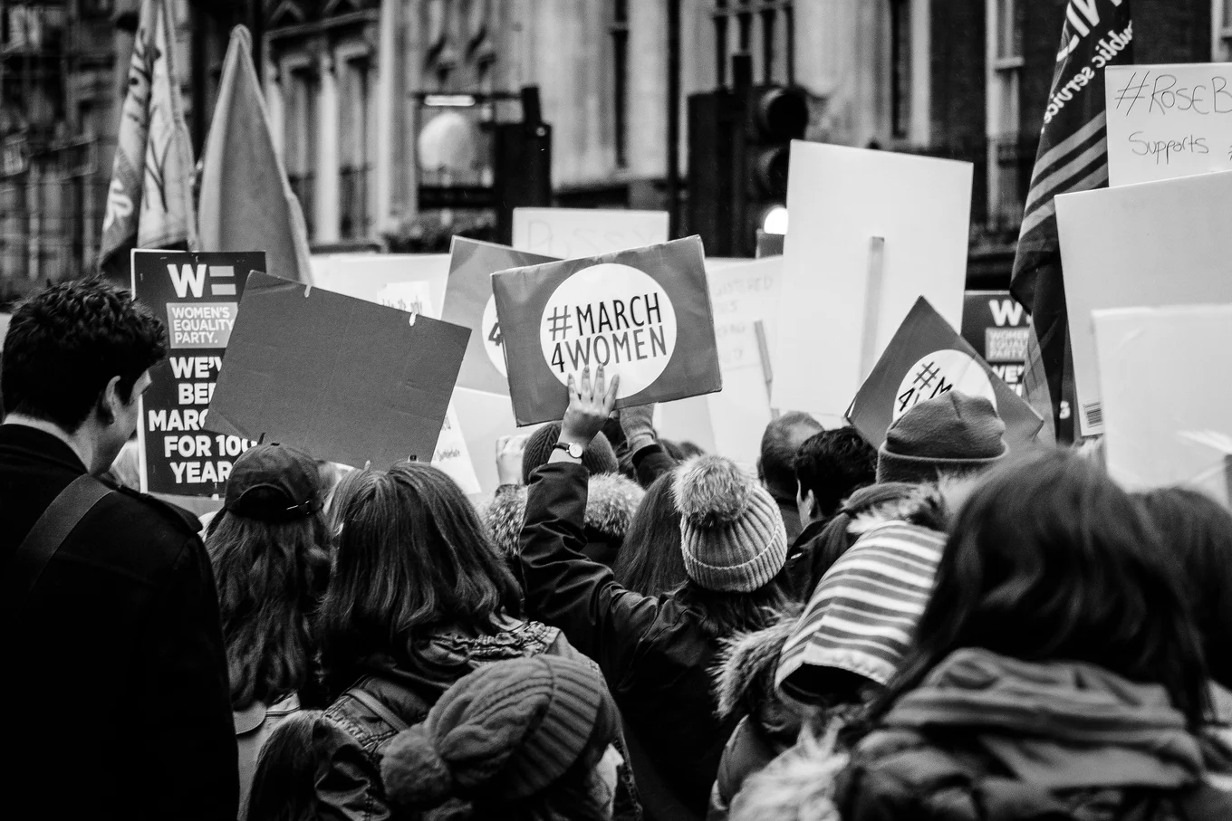 Crowd holding protest signs, one reads "#MARCH 4WOMEN".